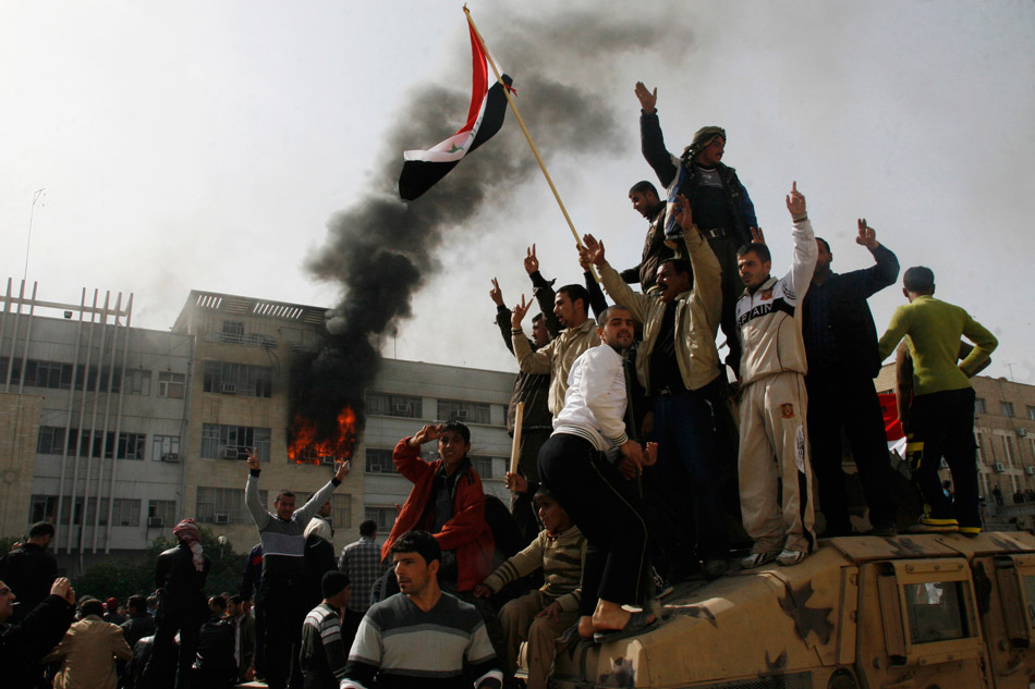 Багдад (Ирак). Демонстранты взбираются на броневик. Виден дым, поднимающийся над зданием администрации губернаторства Мосул во время массовой акции протеста в городе Мосуле, расположенном в 390 км к северу от Багдада. 25 февраля 2011 года. Тысячи иракцев вслед за жителями многих арабских стран вышли на улицы на общенациональный День гнева для того, чтобы выразить протест против коррупции и отсутствия элементарных жизненных условий в стране